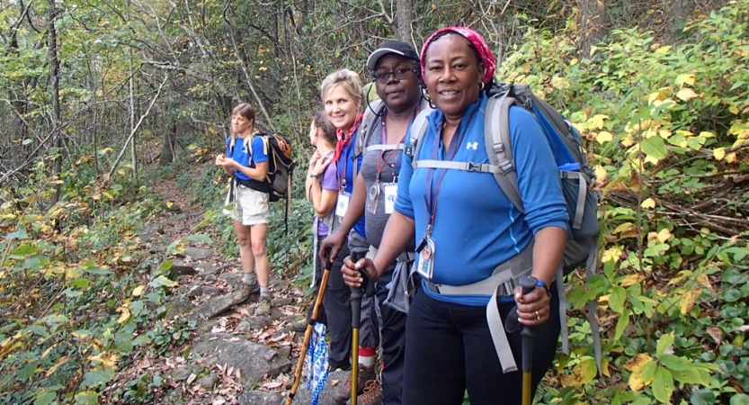 A group of people wearing backpacks stand in a line in a wooded area and smile for the photo